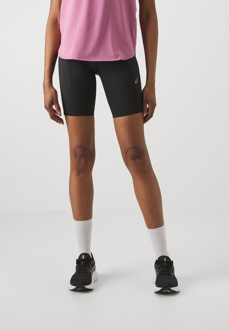 Personne portant un short de sport noir Asics, un haut rose, des chaussettes blanches et des chaussures de course noires, debout sur un sol blanc.