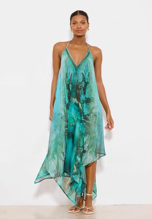 Kvinde stående iført en flydende teal halterneck-kjole med abstrakt mønster og hvide sandaler med stropper mod en ensfarvet hvid baggrund.