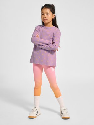 Dziewczyna z długimi ciemnymi włosami stoi z założonymi rękami, ubrana w fioletową koszulkę z długim rękawem z żółtymi wzorami oraz legginsy z gradientem od różowego do pomarańczowego.
