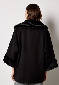 Schwarzer Mantel mit einem weiten, oversized Design. Verfügt über einen Kunstpelzkragen und -manschetten, glattes Material und eine lockere Passform.