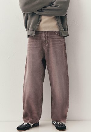 Persona che indossa jeans marroni sbiaditi a gamba larga, sneaker nere con lacci bianchi, una maglia beige e una giacca grigia con zip, con le braccia incrociate.