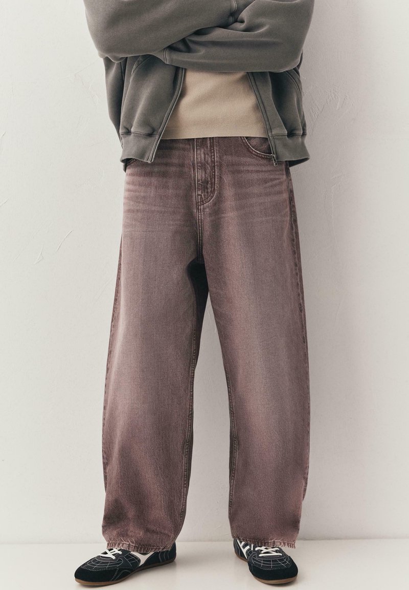 Personne portant un jean large marron délavé, des baskets noires à lacets blancs, un haut beige et une veste grise zippée, les bras croisés.
