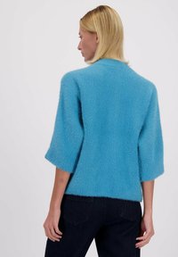 Femme blonde portant un pull en peluche bleu vif avec des manches jusqu'aux coudes et un jean noir, vue de dos sur un fond uni.