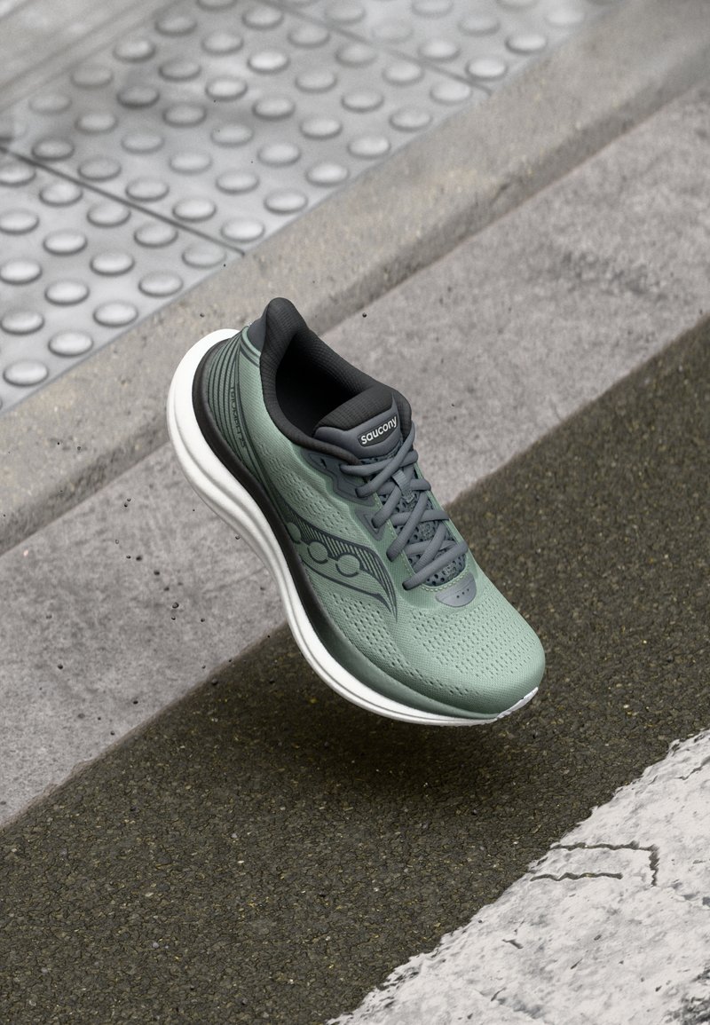 Grønn og svart Saucony løpesko hengt opp over teksturert fortausdekke nær fortauskant og taktilt ledelinjestyringssystem.