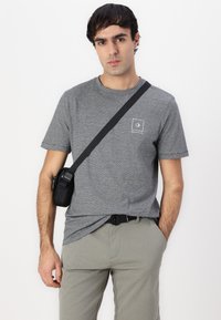 T-shirt gris chiné à manches courtes, arborant un petit logo carré blanc. Associé à un pantalon gris clair et un sac bandoulière noir.