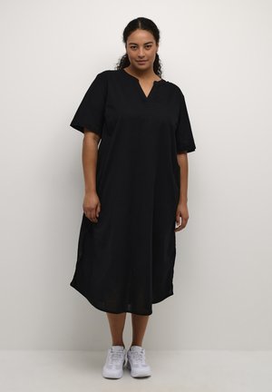 Vestido negro de longitud hasta la rodilla, con mangas cortas, escote en V, aberturas laterales y un corte holgado, hecho de una tela ligera. Combinado con zapatos blancos.