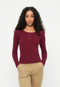 Haut bordeaux à manches longues avec texture côtelée, encolure ronde et fermeture à trois boutons à l'avant. Associé à un pantalon beige.