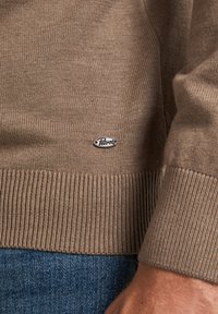 Pull en maille marron avec des poignets et un ourlet côtelés, arborant une petite étiquette de marque métallique à l'avant. Porté par-dessus un jean bleu.