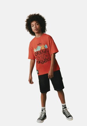 Young person with curly hair wearing an orange Pokémon T-shirt, black shorts, striped socks, and black sneakers standing against white background.