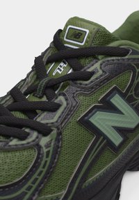 Πράσινο και μαύρο αθλητικό παπούτσι New Balance με μαύρα κορδόνια, επάνω μέρος από πλέγμα και υφή λογότυπο "N" στην πλευρά κοντά στα δάχτυλα και τη γλώσσα.