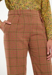 Un pantalon en tissu pied-de-poule rose et marron, taille haute, avec des plis et des poches latérales, associé à une chemise à fleurs à manches longues.