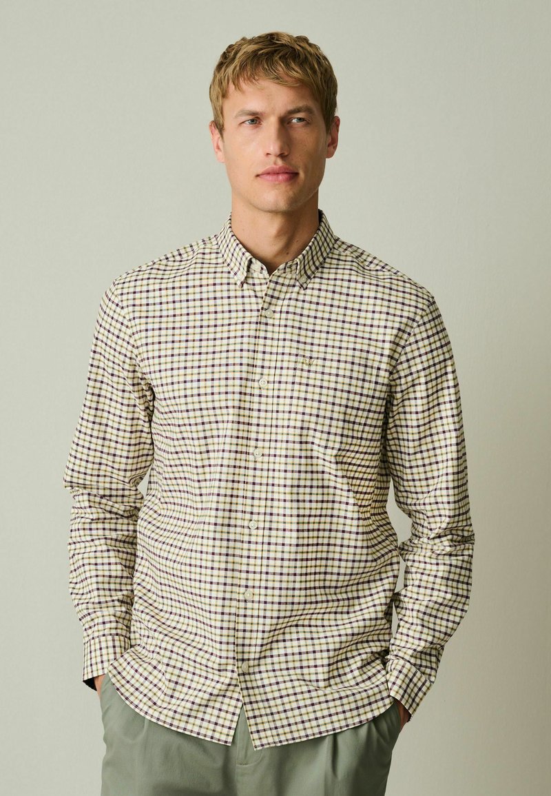 Homme aux cheveux blonds courts portant une chemise beige et marron à carreaux boutonnée et un pantalon vert, les mains dans les poches.