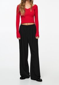 Žena v červeném dlouhém rukávu, zkráceném ribbovaném topu, s černými širokými kalhotami a černými plochými botami, stojící na jednobarevném pozadí.
