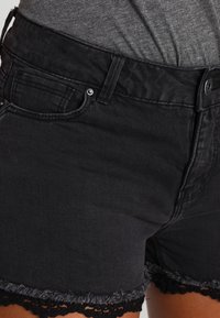 Pantalones cortos de mezclilla negra con un dobladillo deshilachado y adornos de encaje negro. Cuenta con un cierre de botón y cinco bolsillos. Corte regular y textura suave.