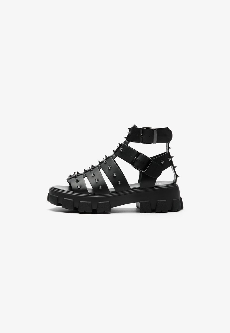Black strappy sandals featuring spiked accents, thick rubber sole, and adjustable buckles. Made of synthetic leather with open design.