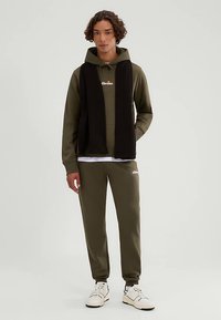 Groene katoenen hoodie met trekkoord, gecombineerd met bijpassende joggers en een zwarte sjaal. Witte sneakers maken de look compleet.