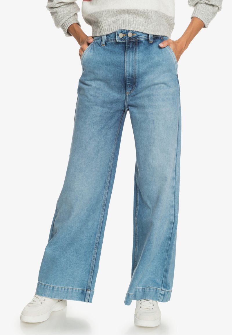 Personne portant un jean taille haute à jambes larges de couleur bleu clair avec une fermeture à double bouton, mains dans les poches, associée à des baskets blanches et un pull gris.
