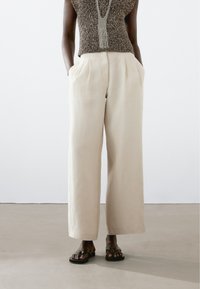 Lichtbeige wijde linnen broek met zijzakken, voorzien van een hoge taille en een zachte textuur. Gesteund met donkere platte sandalen.