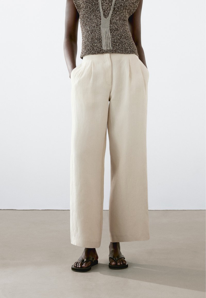 Lichtbeige wijde linnen broek met zijzakken, voorzien van een hoge taille en een zachte textuur. Gesteund met donkere platte sandalen.