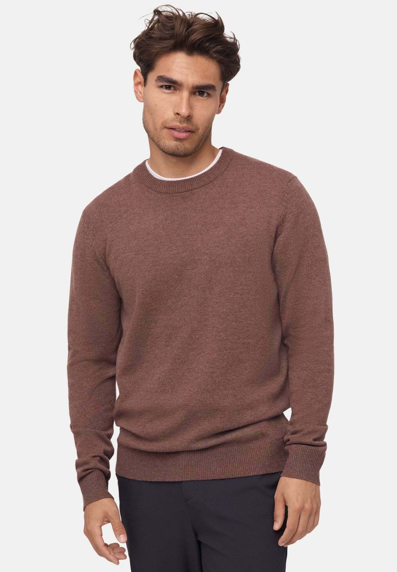 Brązowy sweter z dzianiny z okrągłym dekoltem, ściągaczami na mankietach i u dołu; długie rękawy; miękka tekstura; noszony na białej koszuli.
