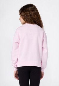 Chica con cabello largo y rizado, vestida con un sudadera rosa claro y pantalones negros, de pie de espaldas contra un fondo blanco.