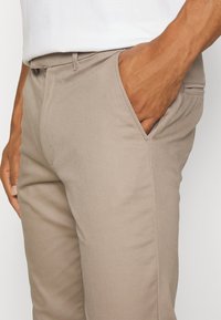 Man med beige byxor och vit skjorta, höger hand i framfickan, nära bild på midjan och övre benen.