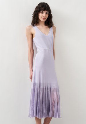 Naiste krussis tumedate juustega, kes kannab varrukateta helerohelist lilla kleiti volditud seelikuga, seisab ühevärvilise valge tausta ees.