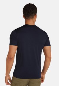 Man med kort lockigt hår som bär en enfärgad navyblå t-shirt med kort ärm och olivgröna byxor, visad bakifrån.