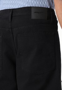 Black denim pants featuring a smooth texture, two back pockets, and a rectangular leather patch with "JACK & JONES" embossed.