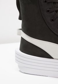 Svart mockaskor med vita läderdetaljer, med en tjock vit sula och platta snören, designade med en rundad topp och minimal sömnad.