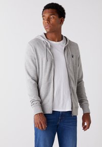 Szara bluza z kapturem na zamek błyskawiczny z troczkami, ściągaczami na mankietach oraz kieszeniami z przodu, noszona na białej koszulce i zestawiona z niebieskimi dżinsami.