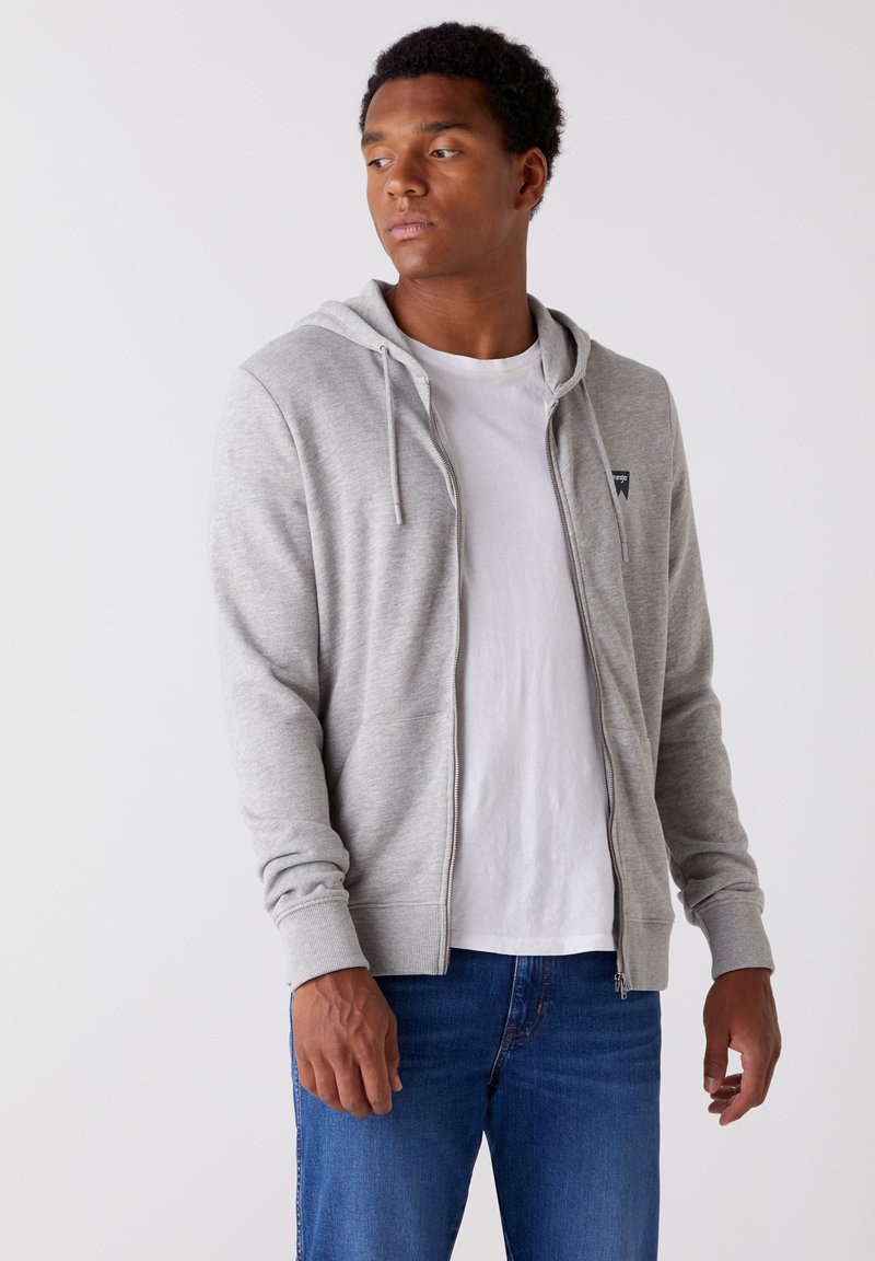 Szara bluza z kapturem na zamek błyskawiczny z troczkami, ściągaczami na mankietach oraz kieszeniami z przodu, noszona na białej koszulce i zestawiona z niebieskimi dżinsami.