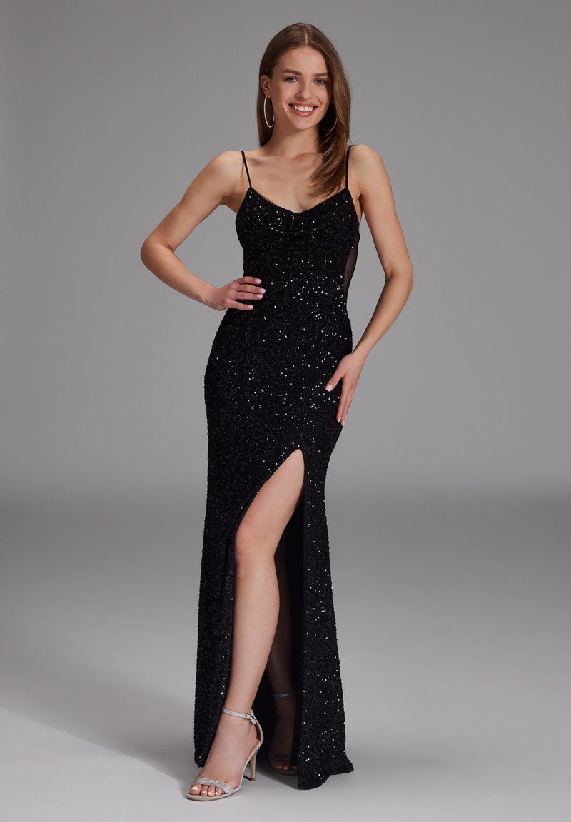 Abito da sera nero con paillettes, dalla silhouette aderente, spalline a spaghetti, alto spacco laterale e pannelli laterali trasparenti, abbinato a tacchi argento.