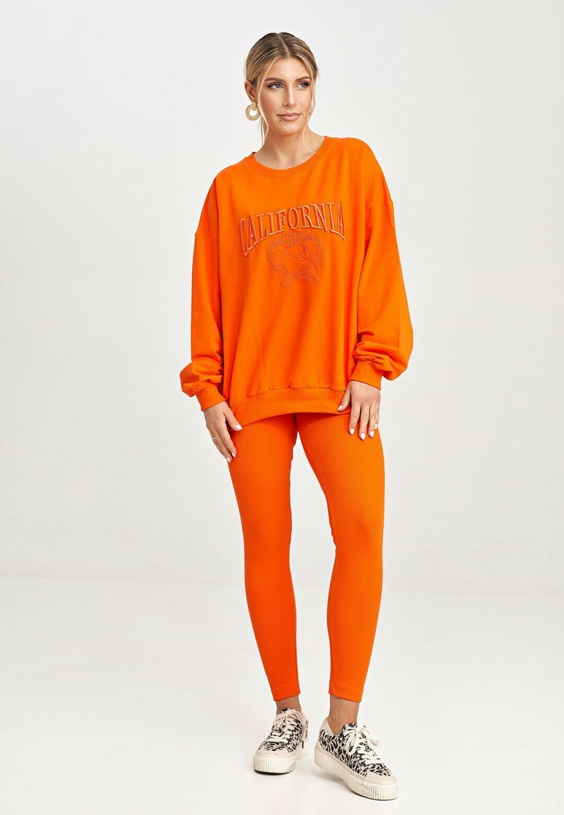 Jasnopomarańczowa, oversize'owa bluza z napisem "CALIFORNIA", w zestawie z dopasowanymi, prążkowanymi legginsami i wzorzystymi sneakersami.