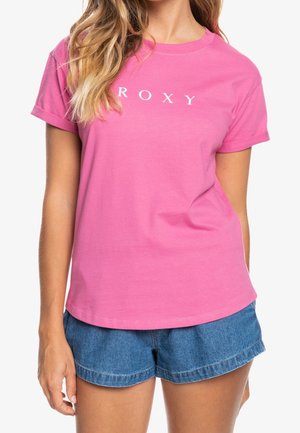 Mujer con camiseta rosa de manga corta con el texto "ROXY" y pantalones cortos de mezclilla azul, posando frente a un fondo blanco.