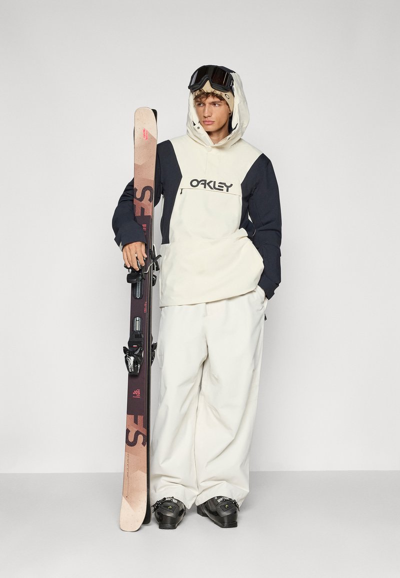 Équipement de ski avec une veste crème et marine, un pantalon blanc et des bottes noires. Tenant un ski sombre avec fixations, portant des lunettes de ski et une capuche.