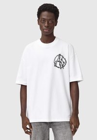 Valkoinen ylipitkä t-paita, joka on valmistettu puuvillasta. T-paidassa on musta graafinen kuvio vasemmassa rinnassa sekä pyöreä pääntie ja lyhyet hihat.