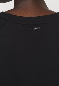 Schwarzer Sweatshirt mit geripptem Halsausschnitt. Verfügt über ein kleines metallisches Logoetikett "DKNY" in der Nähe des Kragens auf einer glatten Stoffoberfläche.