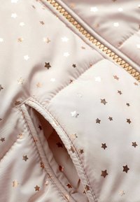 Chaqueta acolchada rosa con patrones de estrellas en oro y cobre, que presenta una cremallera visible y una textura suave. Costuras detalladas y bolsillo visibles.