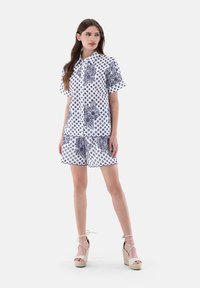 Set di camicia a maniche corte e pantaloni corti in tessuto bianco con motivi floreali e pois blu, caratterizzato da un orlo scallop e chiusura con bottoni.