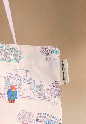 Piros kalapos és kék kabátos Paddington Bear rajzolt mintája, fák, egy kutya, egy busz és épületek mellett egy krémszínű vászontáskán, vászon fogóval.