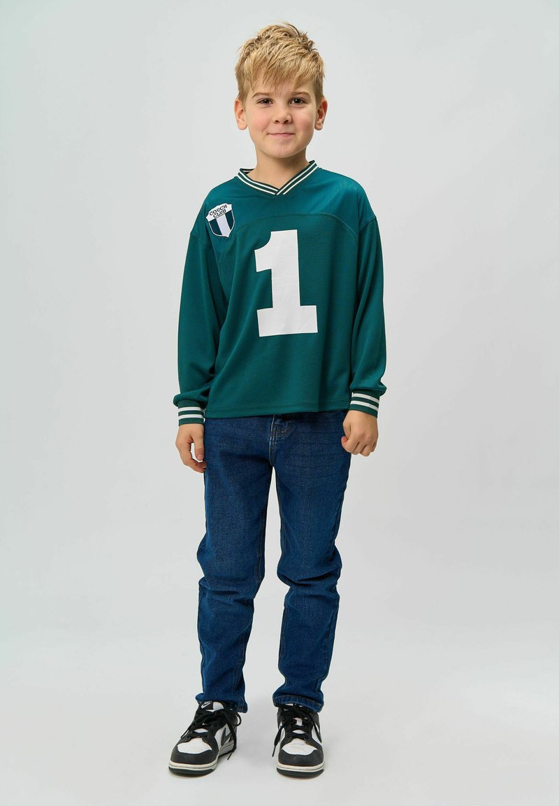 Tricou sportiv verde cu numărul alb "1" și un mic logo, purtat cu blugi bleu și adidași negri cu accente albe.