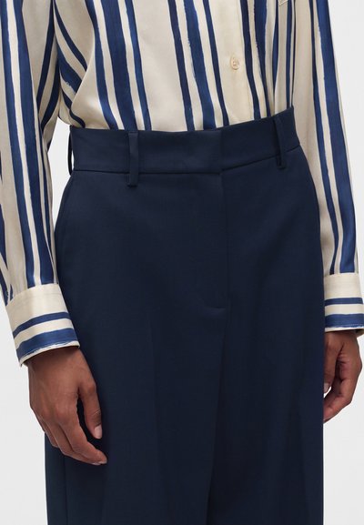 Personne portant un pantalon taille haute bleu marine et une chemise crème à manches longues avec des rayures verticales bleu marine, montrant la taille et les mains.