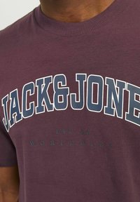 Camiseta de algodón burdeos con cuello redondo acanalado, que presenta "JACK&JONE" en letras grandes blancas y marinas, y un texto de marca en la parte inferior.