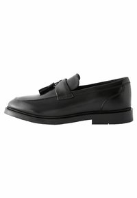 Chaussure noire en cuir à enfiler avec un bout rond, détails cousus et un gland décoratif sur la sangle. Semelle en caoutchouc plate.