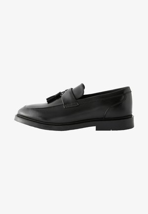 Svart läder slip-on sko med rund tå, sydda detaljer och dekorativ rost på remmen. Plat gummisula.