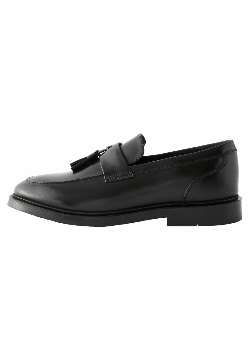 Chaussure noire en cuir à enfiler avec un bout rond, détails cousus et un gland décoratif sur la sangle. Semelle en caoutchouc plate.