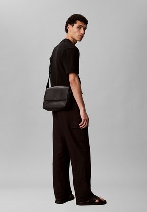 Jeune homme portant une tenue noire et des sandales, avec un sac à bandoulière noir, debout devant un fond gris uni, de profil.