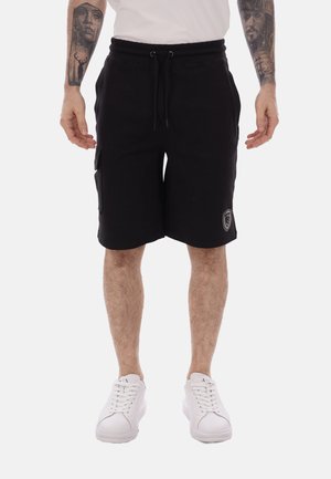 Uomo che indossa pantaloncini neri al ginocchio con tasca laterale e sneaker bianche, con avambracci tatuati visibili, in piedi davanti a uno sfondo bianco.