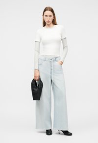 Woman in layered white long- and short-sleeve tops, wide light blue jeans, black heeled boots, holding a black handbag, standing on gray background.